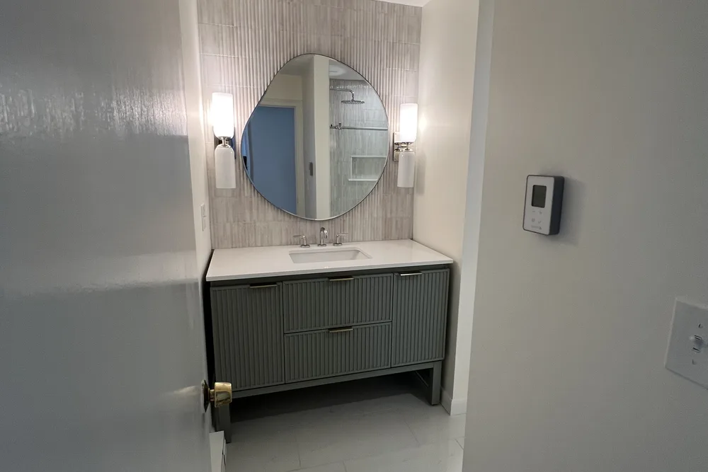 Green vanity with round mirror and wall sconces in a remodeled bathroom by DeFaria Construction