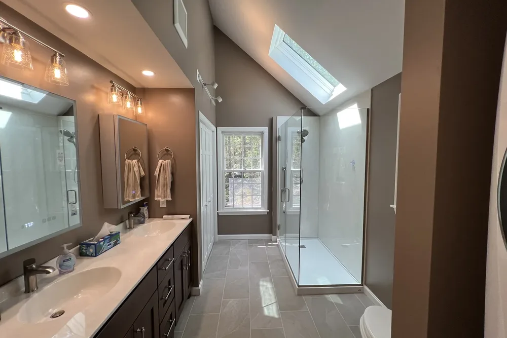 Long primary bathroom with dark vanity, skylight and glass shower by DeFaria Construction